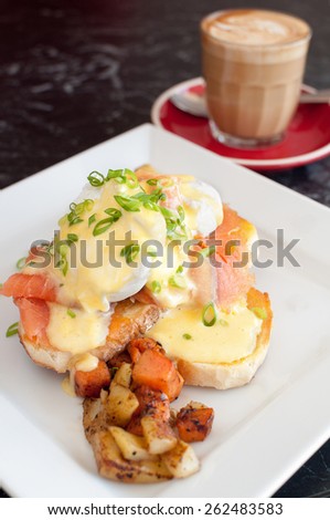 A delicious cooked breakfast of eggs benedict with smoked salmon, toasted sourdough bread and roasted vegetables with a cafÃ?Â© latte coffee in the background