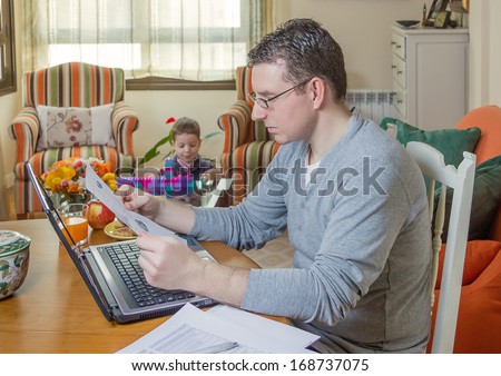 Father reading business documents in home office and his boring son playing on the background