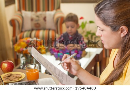 Pregnant mother reading business documents in a home office and his boring son playing on the background