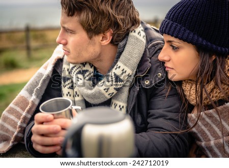 Closeup of young beautiful couple under blanket having hot beverage in a cold day with dark cloudy sky on the background