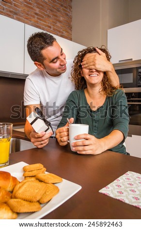 Young man with gift box closing his girlfriend eyes for a surprise while having breakfast on the home kitchen