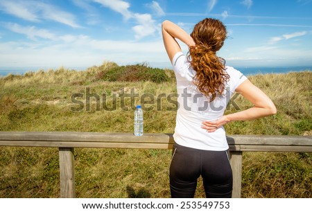 Back view of athletic young woman in sportswear touching her neck and lower back muscles by painful injury, over a nature background. Sport injuries concept.