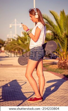 Portrait of beautiful young girl with skateboard and headphones listening music in her smartphone outdoors. Warm tones edition.