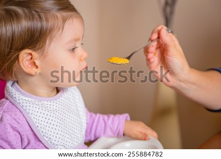 Closeup of adorable happy baby sitting in a feeding chair eating puree from a spoon in the hand of her mother at home