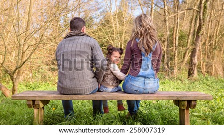 Back view of angry little girl looking to the camera between of man and woman sitting on a wooden bench in the park