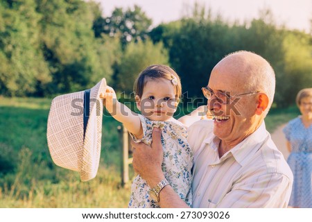 Adorable baby girl playing with the hat of senior man over a nature background. Two different generations concept.