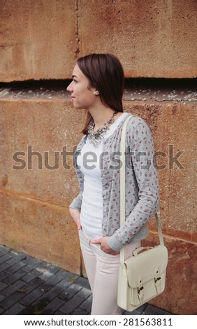 Fashion portrait of young trendy woman wearing gray jacket, pink jeans and white satchel bag over a stone wall background