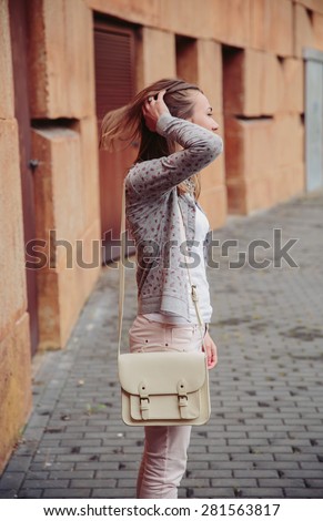 Fashion portrait of young trendy woman wearing gray jacket, pink jeans and white satchel bag over touching her hair outdoors