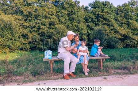 Senior man feeding with fruit puree to adorable baby girl while a jealous brother turns his back sitting in a bench outdoors. Grandparents and grandchildren relations concept.