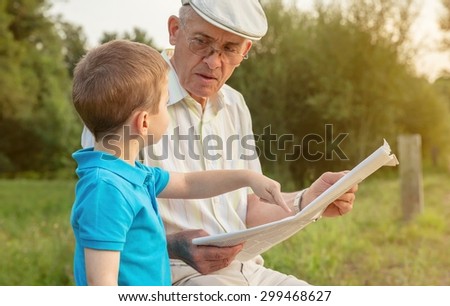 Closeup of senior man reading newspaper and cute child pointing an article with his finger sitting over a nature background. Two different generations concept.