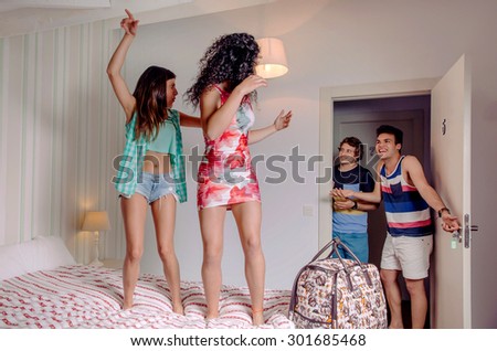 Young happy women couple dancing over a bed and their friends laughing to open the room door. Young people lifestyle concept.