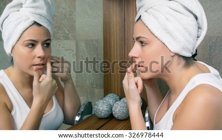 Closeup of young woman with a towel over hair squeezing an acne pimple in her beautiful face in front of a mirror