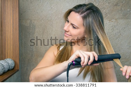Closeup of smiling young woman straightening her hair with a straightener in the bathroom. Health and beauty concept.