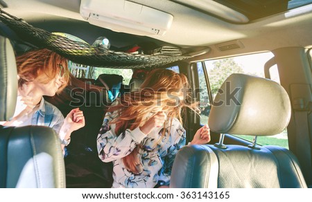 Two happy young women friends dancing and having fun inside of car in a road trip adventure. Female friendship and leisure time concept.