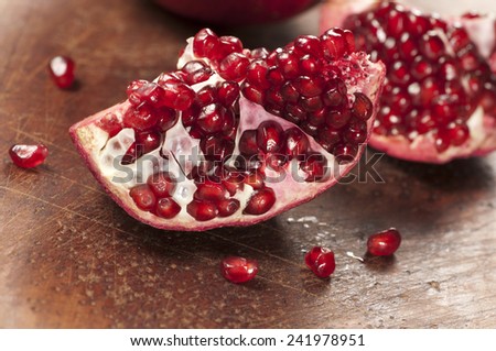 Pomegranate sitting on a dark brown, wooden cutting board. The pomegranate is cut open, with several seeds falling out
