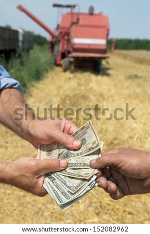 Farmer and buyer hands holding dollar banknote in wheat field with combine in background