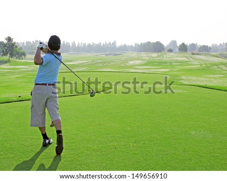 Man golf player with driver teeing-off from tee-box to ward, view from behind.