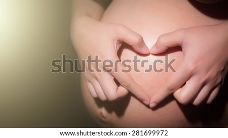 woman holding her hands in a heart shape on her baby bump pregnant belly with fingers on black background