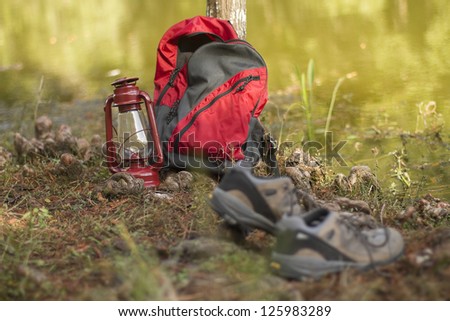 Camping/hiking gear by the side of a placid lake. Shoes,backpack, and lantern.