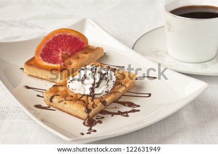 A sweet moment - cup of black coffee and two waffles with whipped cream, chocolate and a slice of red orange on white background
