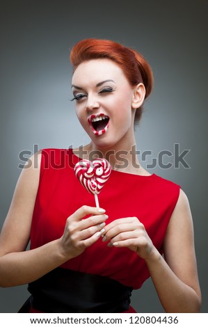 young winking caucasian red-haired woman in red dress holding lollipop with funny expression