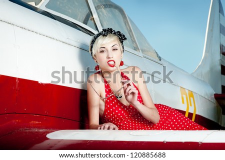 outdoors pin-up portrait of young blond sexy caucasian woman in red retro dress lying on airplane wing