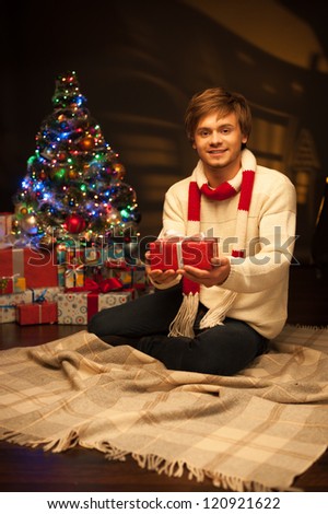 young happy smiling casual man holding red gift over christmas tree and lights on background. warm light