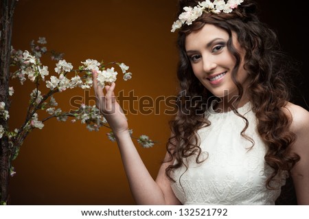 young smiling brunette woman in long white dress and flowers in hair standing near flowering tree over orange background