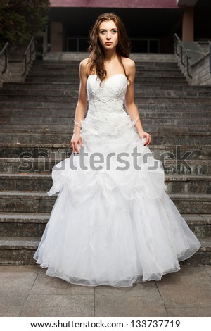 outdoors portrait of beautiful young caucasian brunette woman in white wedding dress over gray stairs on background