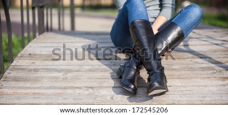 female legs in long black leather boots sitting at wooden bridge