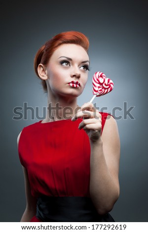 young sexy caucasian red-haired woman in red dress holding lollipop over gray background