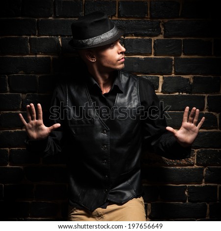 young caucasian man in black shirt and hat lean against black brick wall
