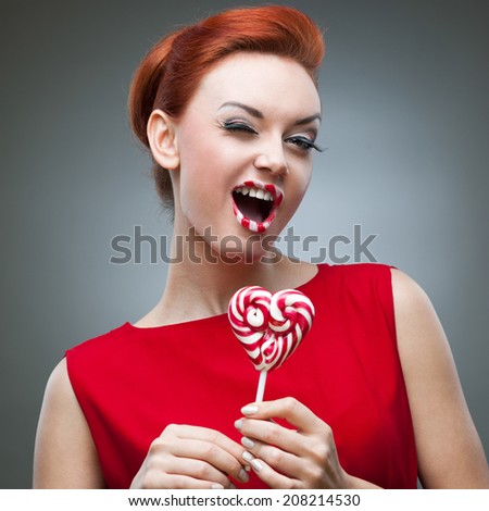 young funny caucasian red-haired woman in red dress holding lollipop over gray background