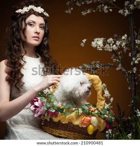 young sensual brunette woman in long white dress and flowers in hair holding basket with puppy  over orange background