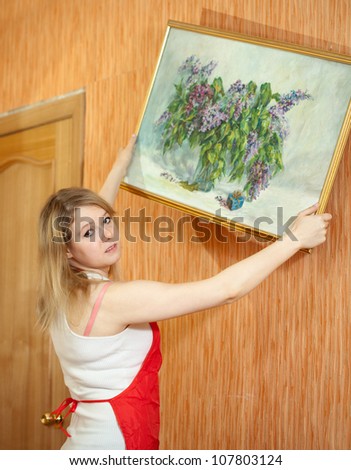 Young woman  hangs the art picture on wall at home