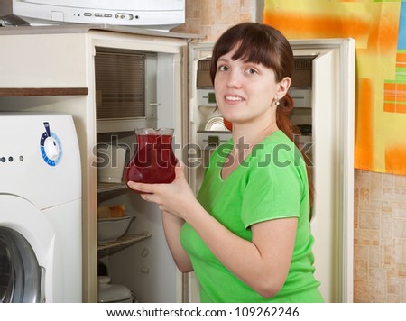 Young woman putting jug with fruit-drink into fridge at home