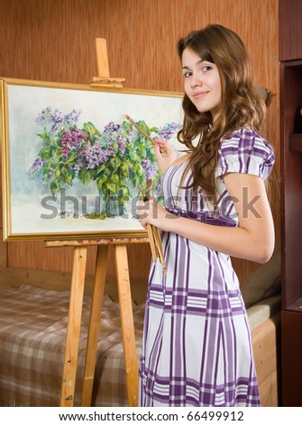 Female painter with brushes near  easel with picture of flowers in interior