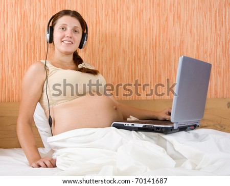 Pregnant woman with headphones and laptop on bed