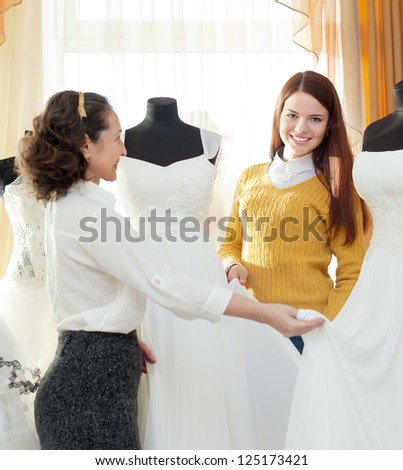 Female shop consultant helps girl chooses white bridal outfit at shop of wedding fashion. Focus on bride
