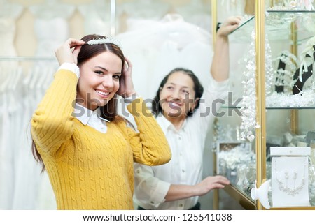 Mature woman helps the bride in choosing bridal diadem at wedding store. Focus on girl