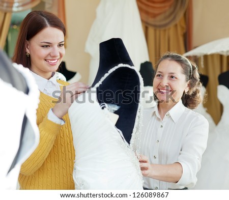 Shop assistant  helps to girl chooses white bridal outfit  at shop of wedding fashion. Focus on bride