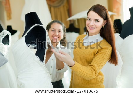 Mature woman helps the bride in choosing bridal gown at wedding store. Focus on girl