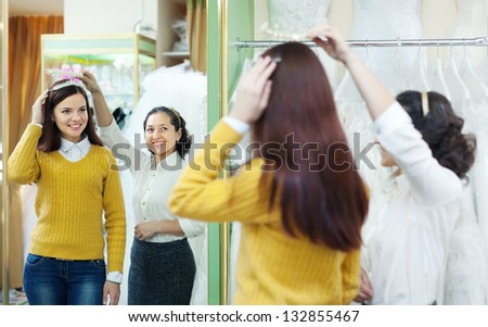Mature saleswoman helps bride chooses bridal accessories at shop of wedding fashion. Focus on reflection