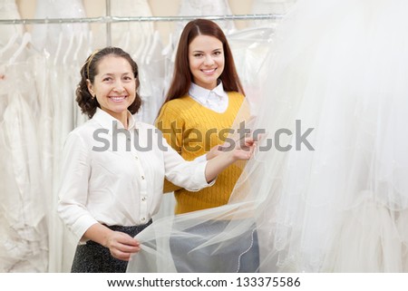 Woman helps the bride in choosing bridal veil at shop of wedding fashion. Focus on mature