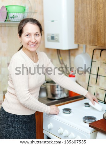 Happy mature housewife cleans the gas-stove  with sponge  in kitchen at home