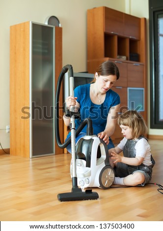 Woman teaches baby girl to use the vacuum cleaner in living room