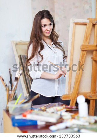 Long-haired girl paints with oil colors on easel in workshop interior