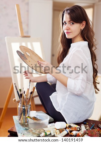 Long-haired female artist with oil colors and brushes near  easel with blank canvas ready for job  in workshop