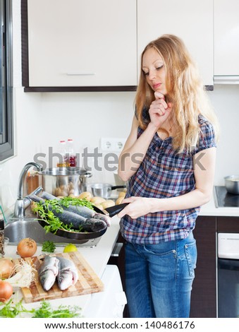 Long-haired woman is thinking how to cook fish in fryingpan at kitchen