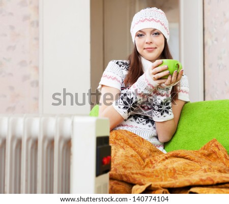 Beautiful woman relaxing near oil heater at home with cup in winter